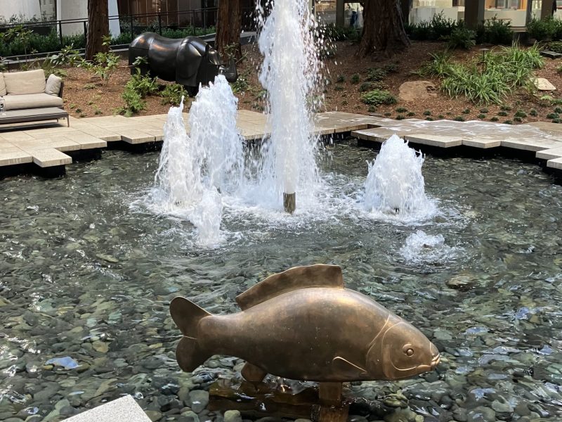 A courtyard fountain surrounded by trees features sculptures of a fish and a rhinoceros. Water jets spray upwards from the fountain, with building structures visible in the background.