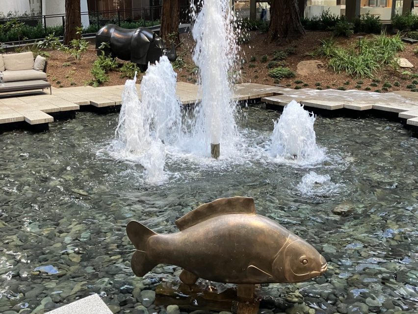 Bronze fish sculpture and water fountains in a stone-lined pond, with a rhino statue and seating area in the background.