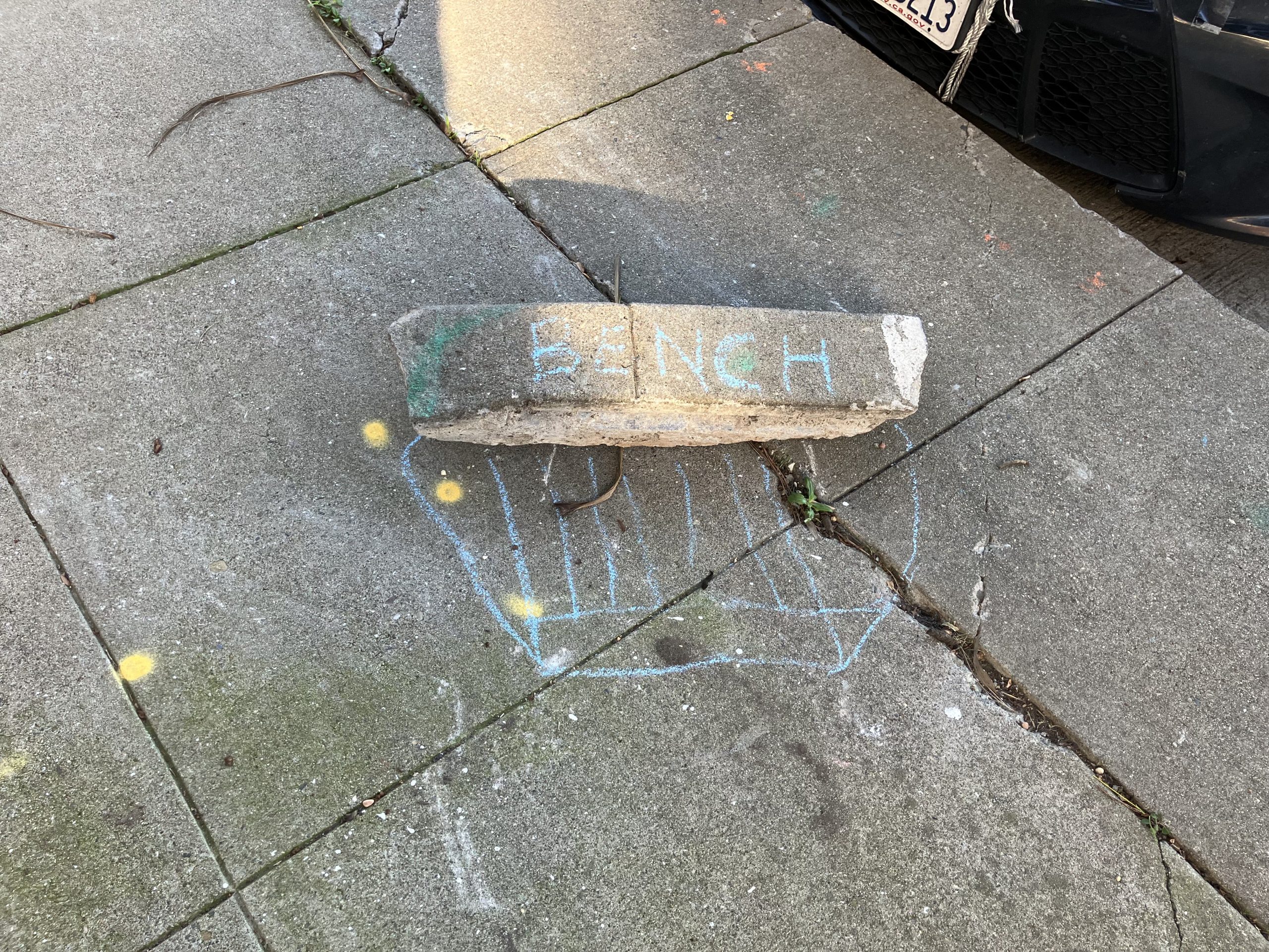 A concrete block labeled "BENCH" on a sidewalk, with a simple chalk drawing resembling a seat outline surrounding it.