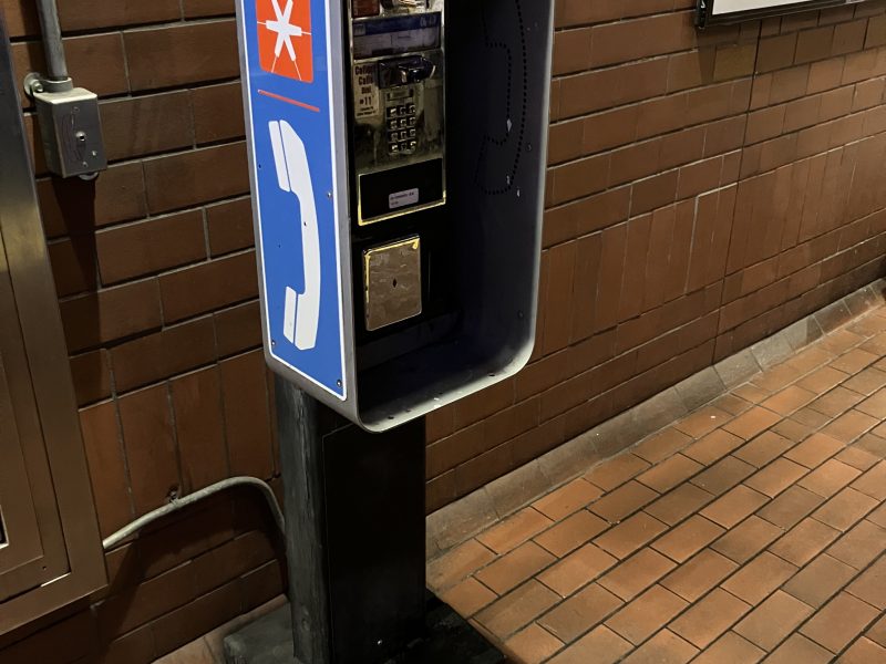 A wall-mounted payphone inside a cubicle on a tiled floor against a brick wall. The phone has a blue sign with a white phone icon and an asterisk symbol in an orange square.