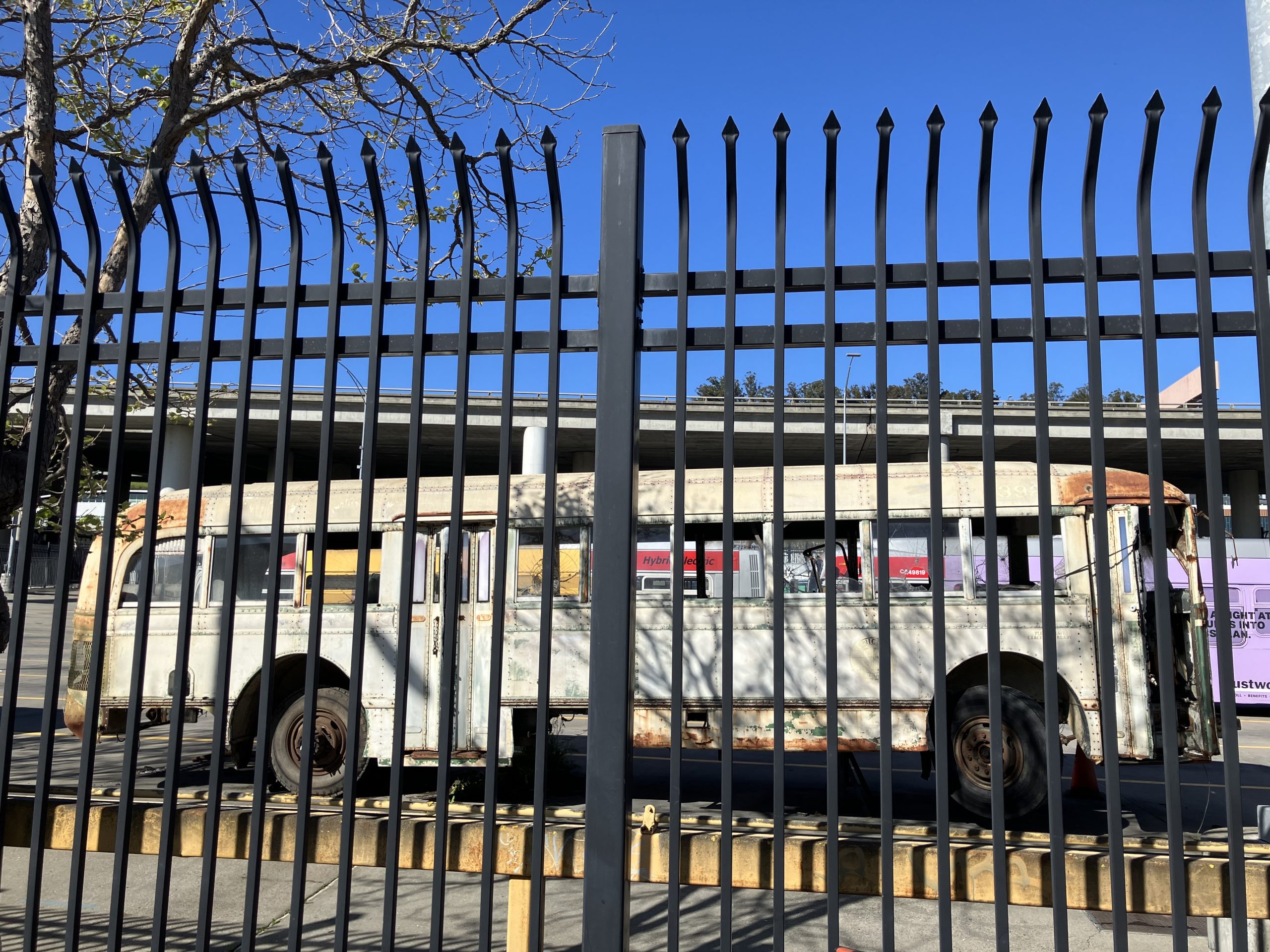 A rusted, vintage bus is parked behind a tall, black metal fence, with a clear blue sky and highway overpass in the background.