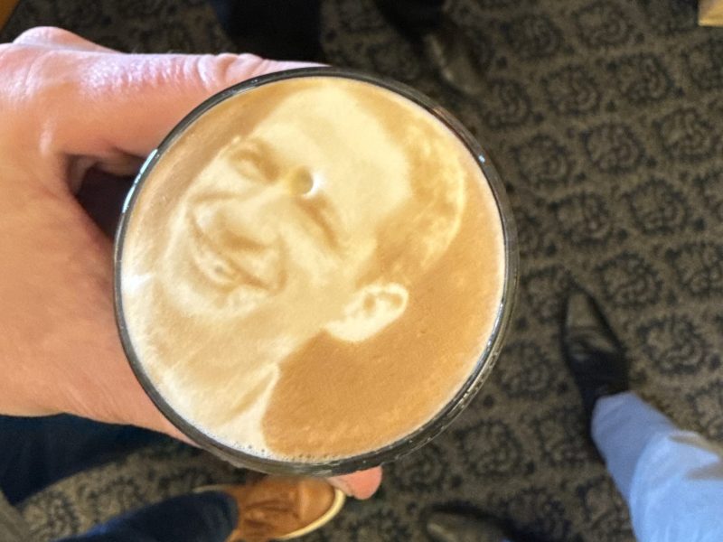 A hand holds a cup with latte art depicting a person's face. Two pairs of feet and the patterned carpet are visible in the background.
