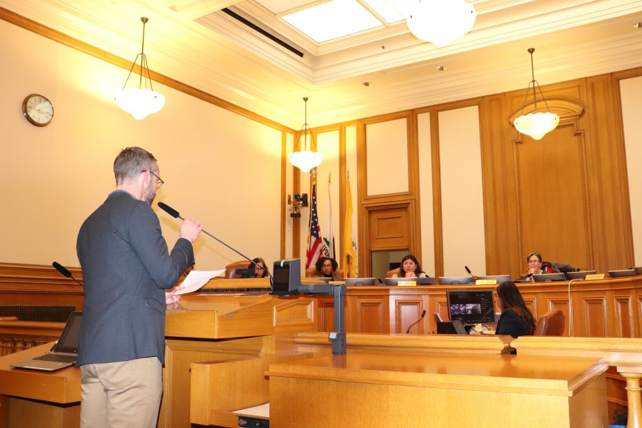 A person speaks at a podium in a courtroom-style setting, addressing a panel of five individuals seated at a raised desk. An American flag and a clock are visible in the background.
