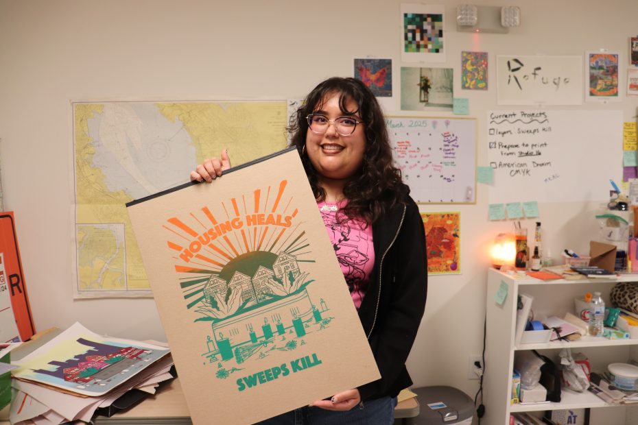 A person smiling and holding a poster titled "Housing Heals, Sweeps Kill," standing in an office with various artworks and papers on the wall and shelf.