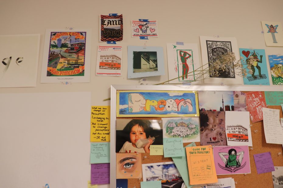A wall collage featuring various posters, postcards, and notes pinned and taped, displaying colorful illustrations, photos, and handwritten messages in an arranged, casual manner.