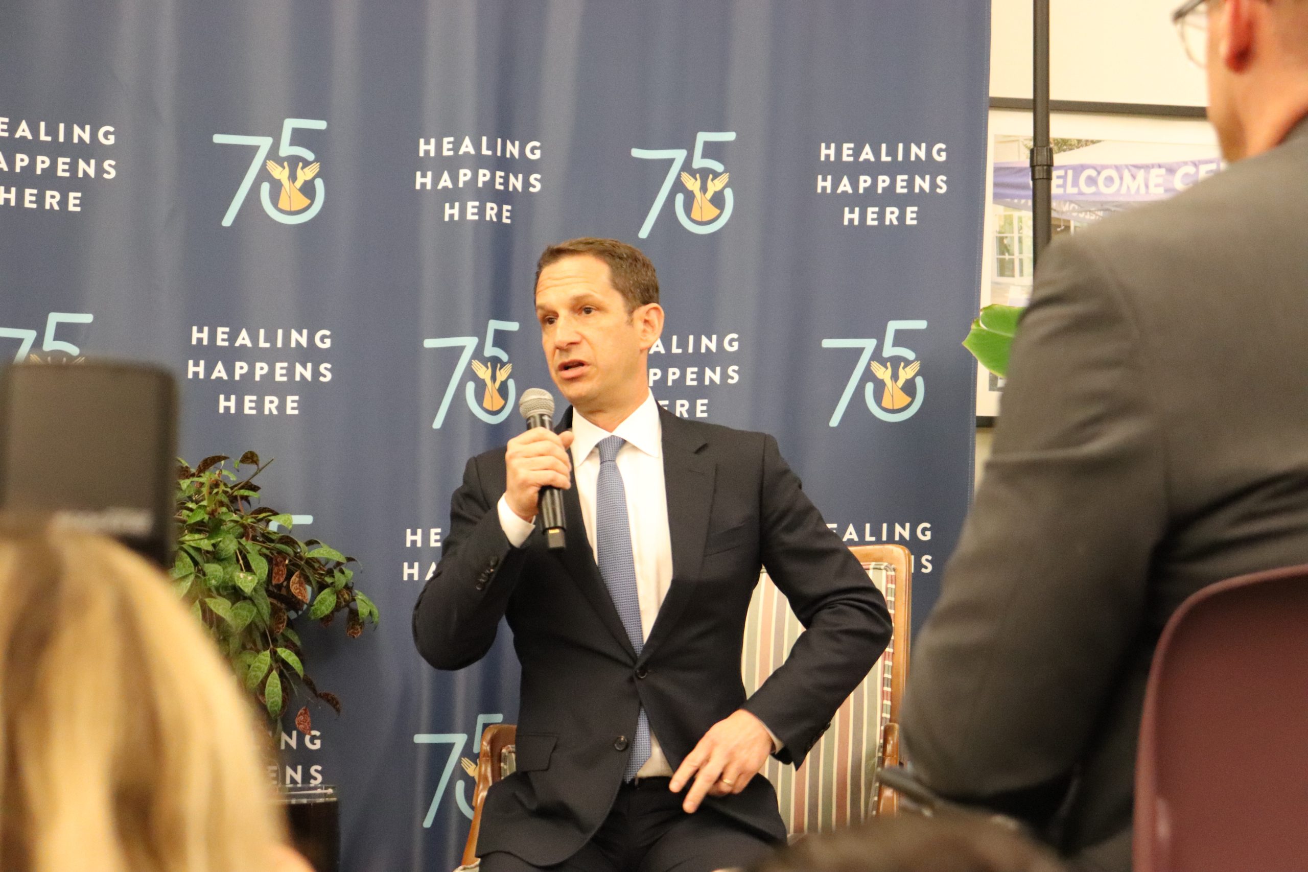 A man in a suit speaks into a microphone at an event. The backdrop displays "Healing Happens Here" and "75" with a logo. Audience members are partially visible in the foreground.