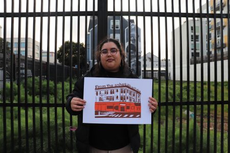 Person holding a sign with a building illustration and the text "From the ashes we will rise" in front of tall metal fencing and urban background.