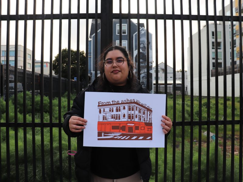 Person holding a sign with a building illustration and the text "From the ashes we will rise" in front of tall metal fencing and urban background.
