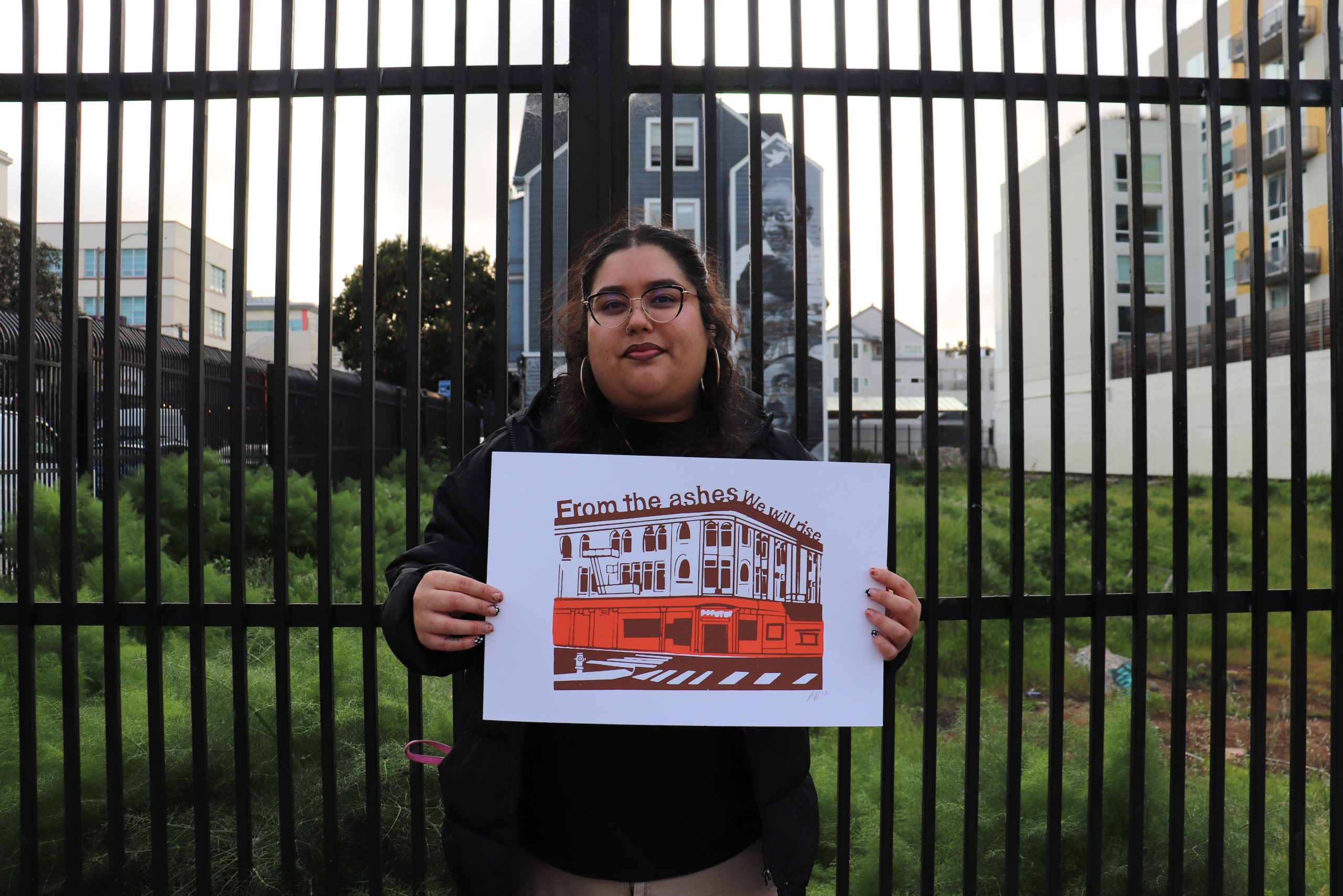 Person holding a sign with a building illustration and the text "From the ashes we will rise" in front of tall metal fencing and urban background.