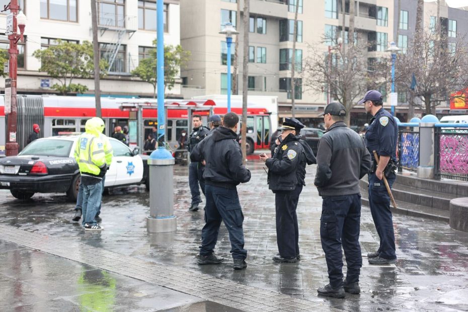 A group of SFPD officers gathers in a wet urban area, surrounded by patrol cars and city buses.