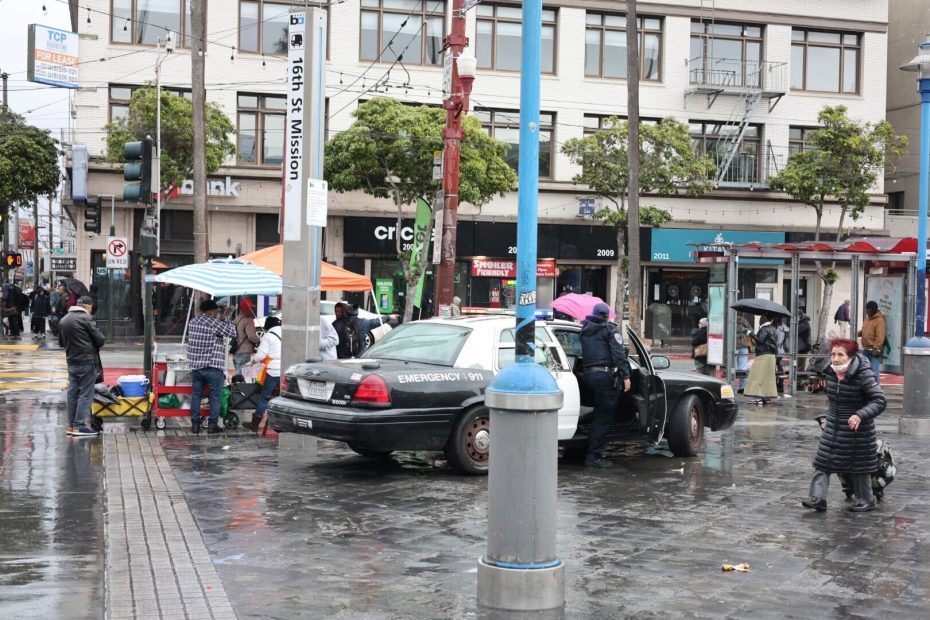 SFPD officers stand vigilant near their car on a rainy street, mingling with pedestrians and tents under the somber sky. Beyond them, businesses hustle in the background while commuters gather at the nearby bus stop, braving the wet weather.