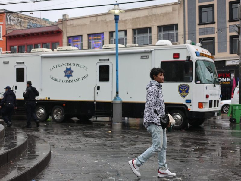 A young man walks in front of an SFPD Mobile Command vehicle parked on a rainy San Francisco street, with officers in the background and buildings surrounding the area.