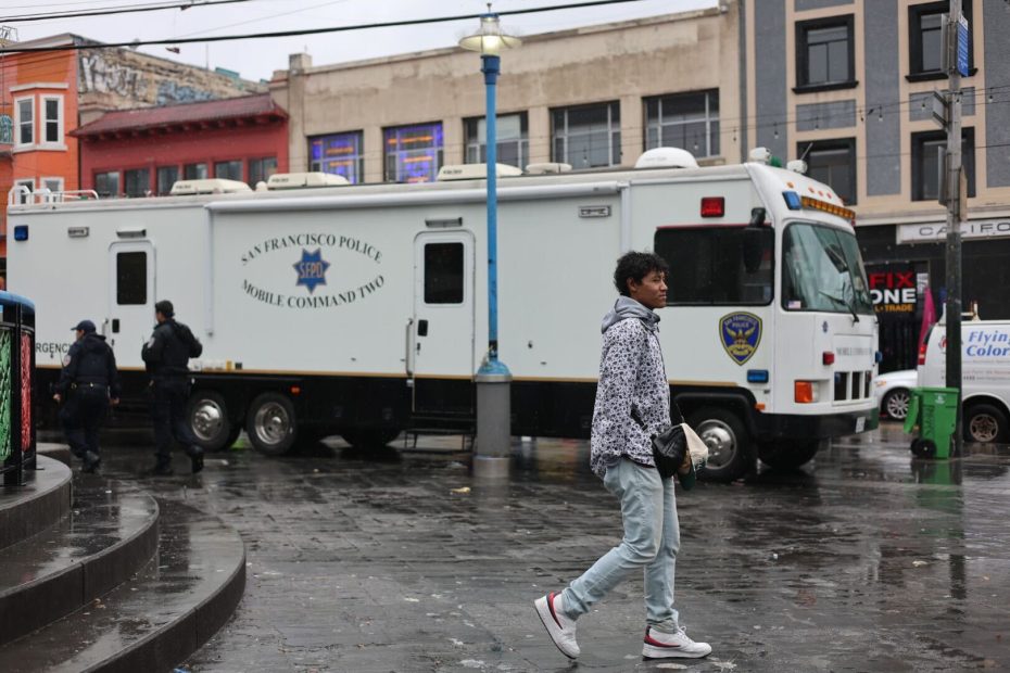 A young man walks in front of an SFPD Mobile Command vehicle parked on a rainy San Francisco street, with officers in the background and buildings surrounding the area.
