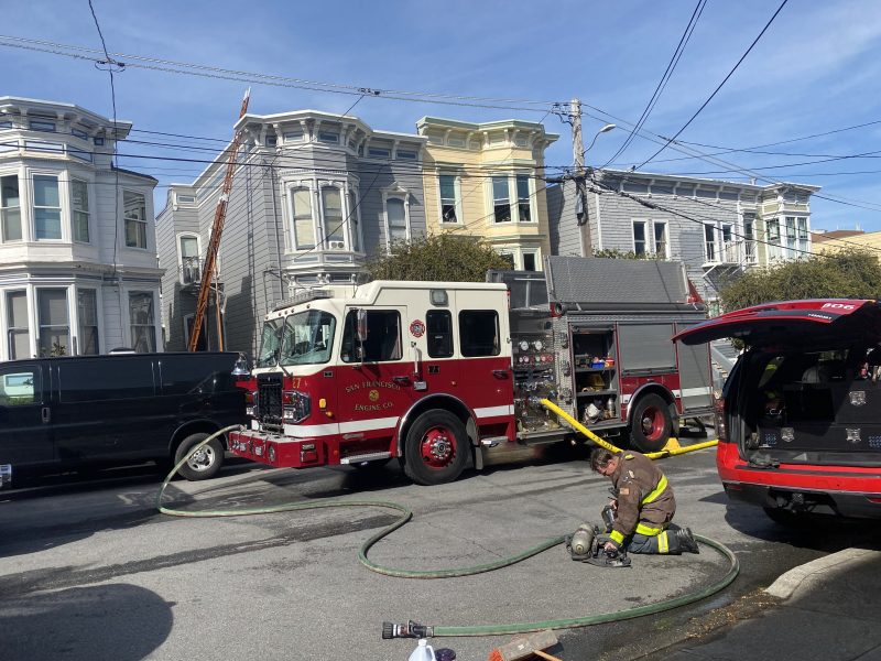 A firefighter kneels next to equipment on the street near a fire truck with hoses extended. A ladder is positioned against a residential building.