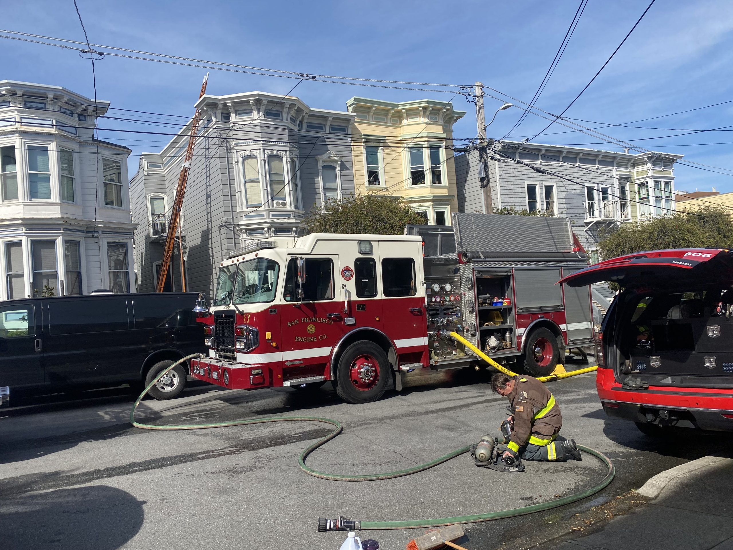 A firefighter kneels next to equipment on the street near a fire truck with hoses extended. A ladder is positioned against a residential building.