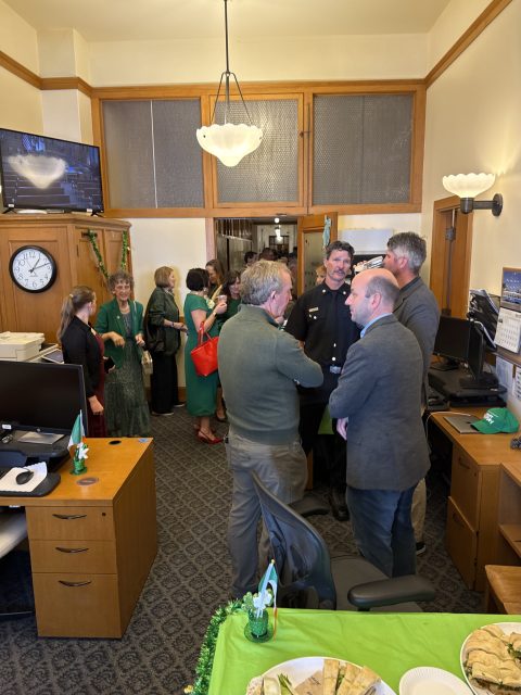 A group of people conversing in an office decorated with green streamers, near desks and a TV. A wall clock shows 10:10. Plates with snacks are on a green table in the foreground.