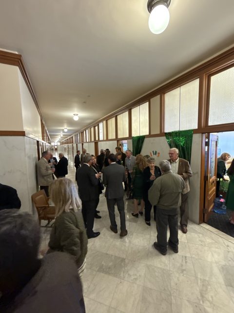 People in business attire mingle in a hallway with marble floors and wood-paneled walls, at a social gathering or event.