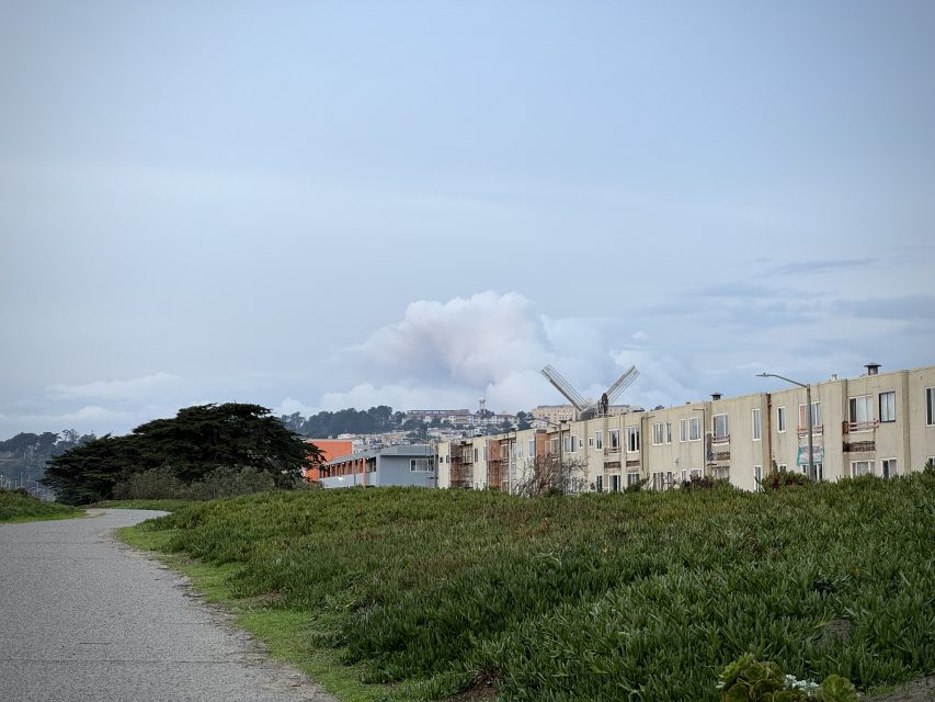 Pathway alongside a grassy field and row of houses under a cloudy sky. Two windmills are visible in the distance.