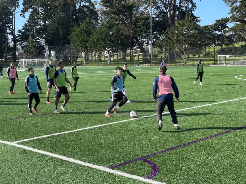 Soccer players scrimmage on a green field under a clear sky, wearing mixed jerseys. Trees and a fence are in the background.