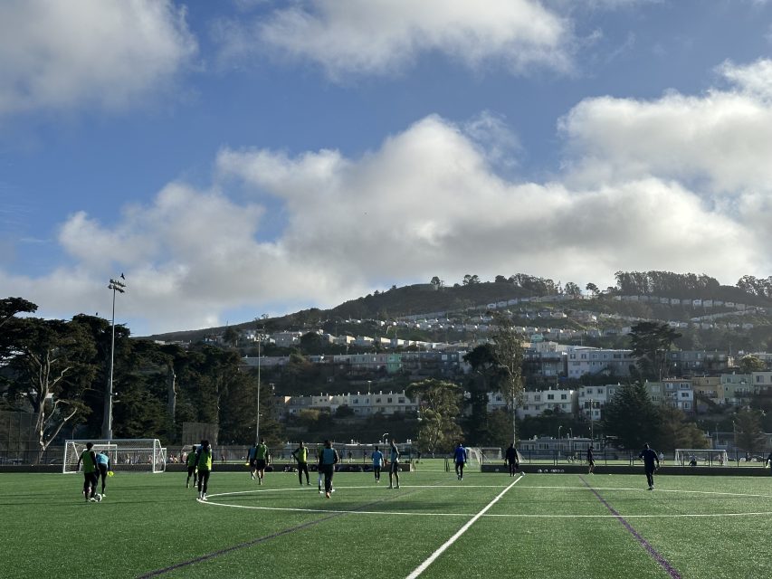 People playing soccer on a field, with several players and goalposts visible. Trees and houses on hills in the background under a cloudy sky.