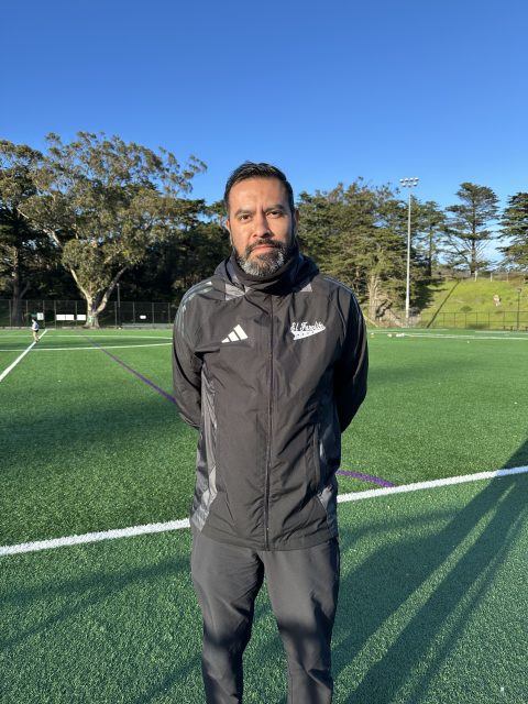 A person stands on a soccer field wearing a black jacket with a logo and Adidas stripes. Trees and a clear sky are in the background.
