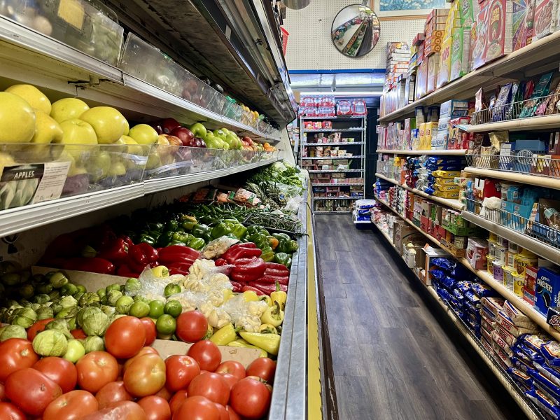 Grocery store aisle with fresh produce like tomatoes, peppers, and lemons on the left, and packaged goods like cereals and snacks on the right. Mirror on ceiling in the background.