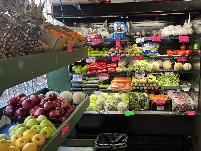 Fruit and vegetable display with apples, pineapples, oranges, cucumbers, carrots, peppers, lettuce, herbs, and other produce on shelves in a market.