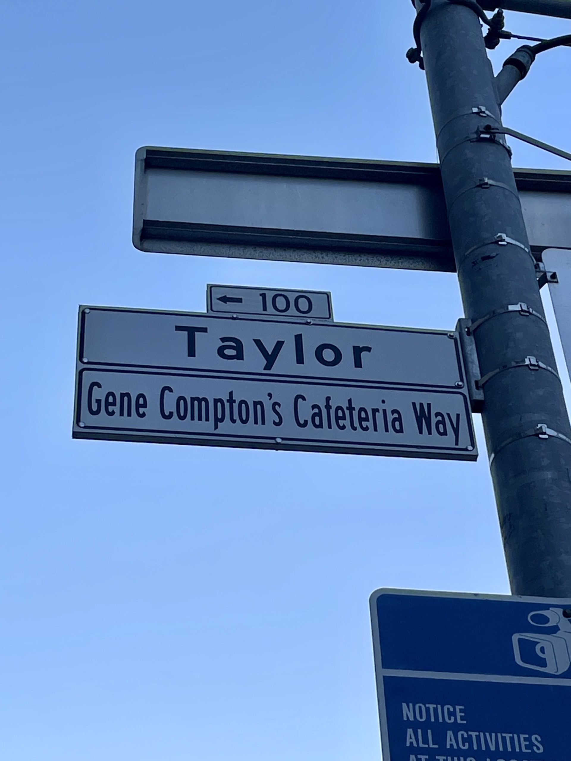 Street signs reading "Taylor" and "Gene Compton's Cafeteria Way" with a directional arrow and a pole against a clear blue sky.