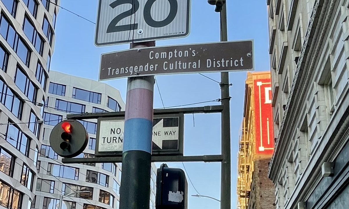 Street signs including a speed limit, Compton's Transgender Cultural District, no left turn, and one way, against a backdrop of buildings and traffic light.
