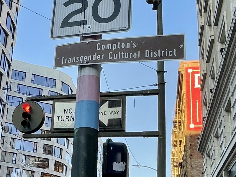 Street signs including a speed limit, Compton's Transgender Cultural District, no left turn, and one way, against a backdrop of buildings and traffic light.