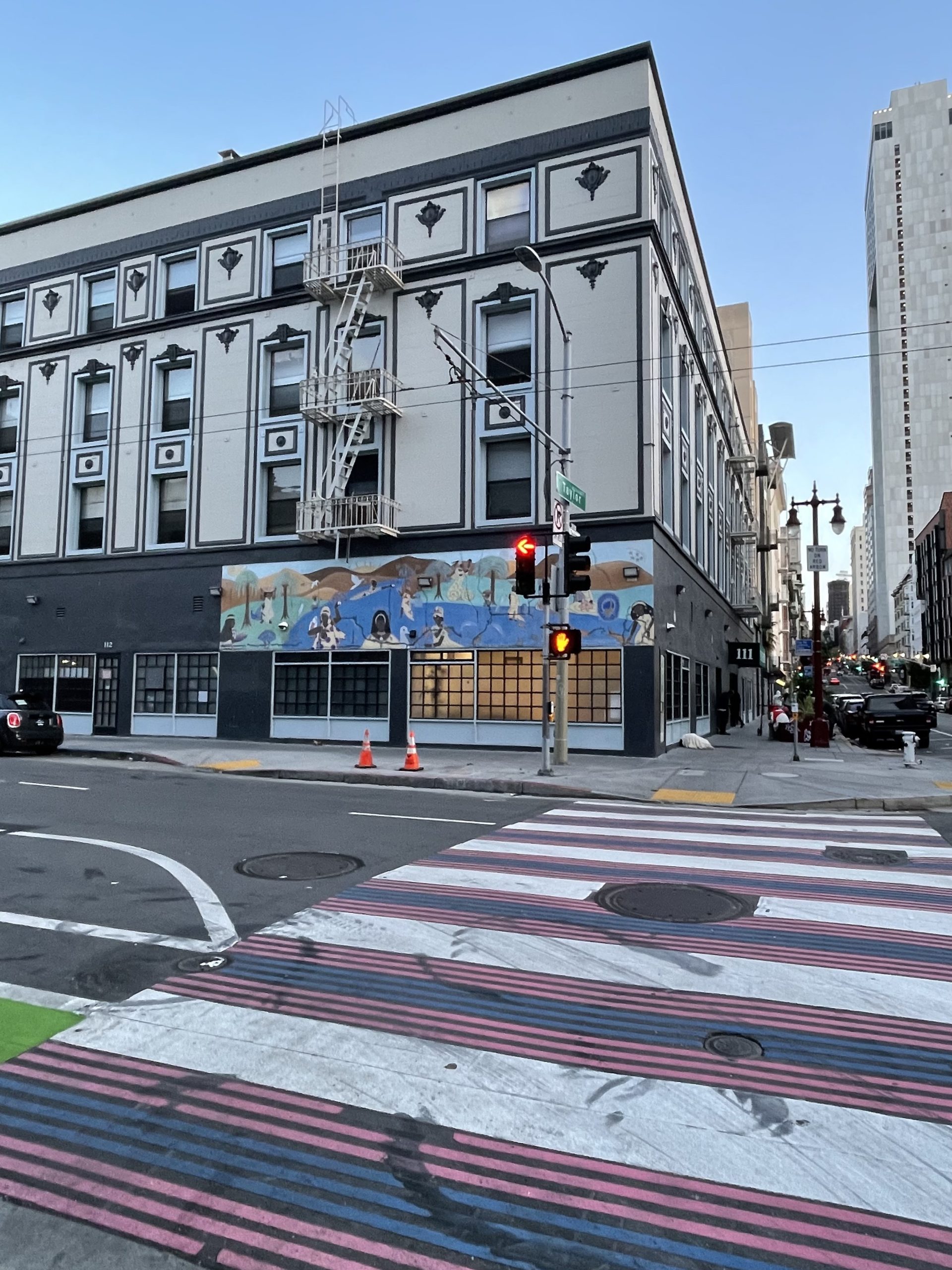 Street corner with a three-story building featuring a mural above the sidewalk. The ground has a colorful crosswalk and traffic lights are present. City buildings are visible in the background.