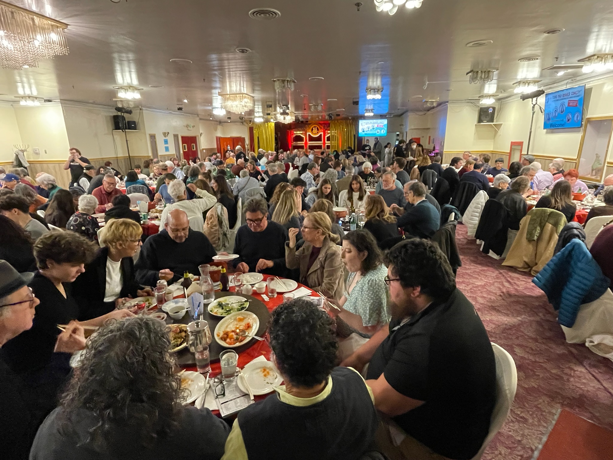 A large group of people are seated at round tables, eating and socializing in a banquet hall with chandeliers and red curtains.