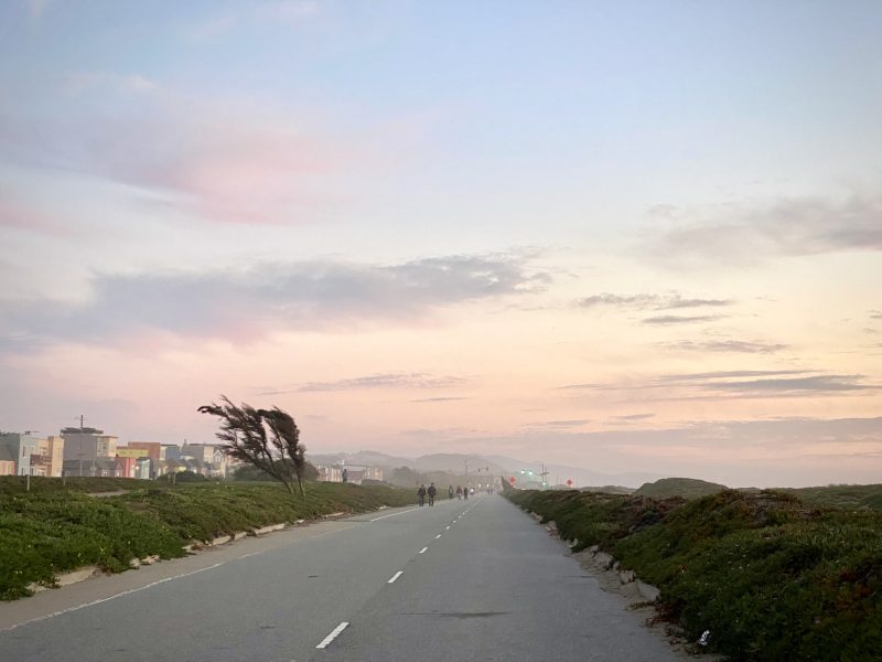 A straight road leads into the distance under a purple and pink sky. Trees on the left are bent by wind, with buildings in the background. People walk along the road.