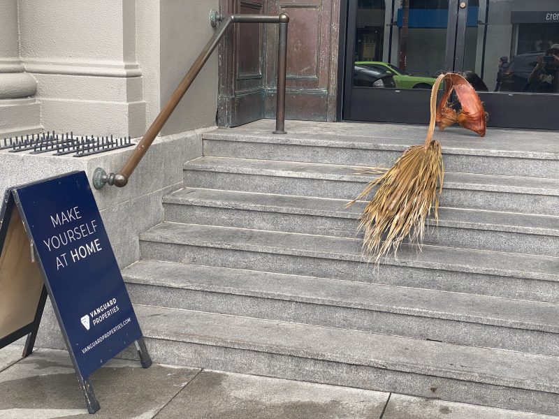 A plant arrangement resembling a large seed pod and straw leaning on steps next to a sign reading "Make Yourself at Home" outside a Vanguard Properties entrance.