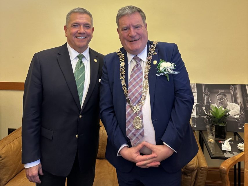 Two men in suits stand smiling indoors. One wears a chain of office and a flower on his lapel.
