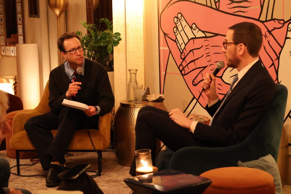 Two men seated and holding microphones are engaged in a conversation. One man is holding a book. A large artwork with interlocking hands is in the background.