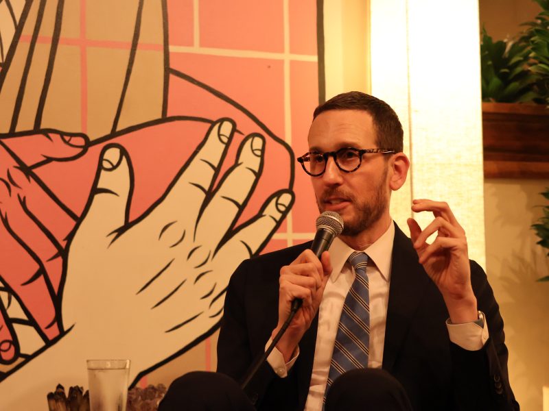 A person in glasses and a suit holds a microphone, speaking in front of a colorful background with large artwork of hands.