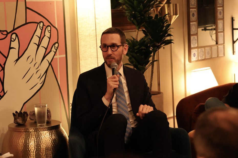 A man wearing glasses and a suit speaks into a microphone while seated in a warmly lit room with plants and artwork in the background.