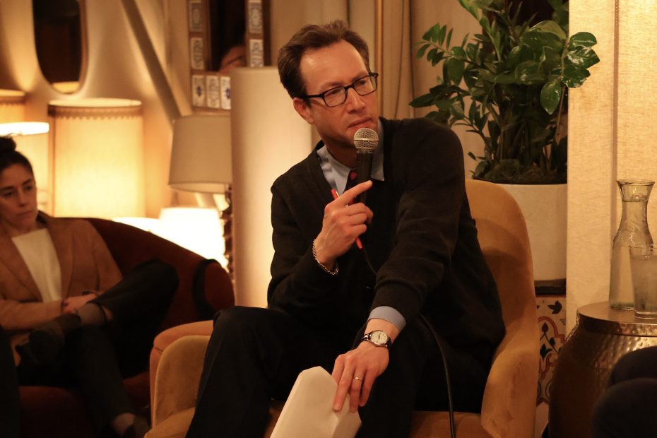 Man in glasses holding a microphone, seated and pointing with a pen in hand. Another person listens in the background. Indoor setting with soft lighting and plants.