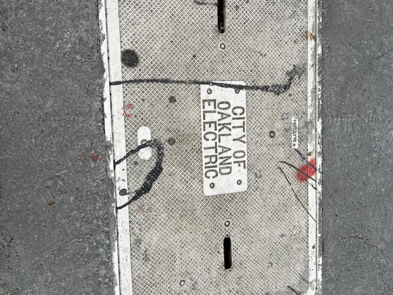 Rectangular manhole cover labeled "City of Oakland Electric" on a concrete sidewalk, with scattered black and red markings.