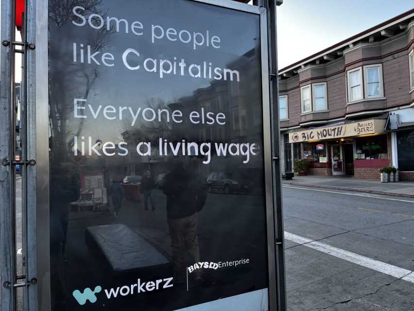 Bus stop advertisement reads, Some people like Capitalism, Everyone else likes a living wage, with logos for workerz and BaySideEnterprise. Background shows a city street and buildings.