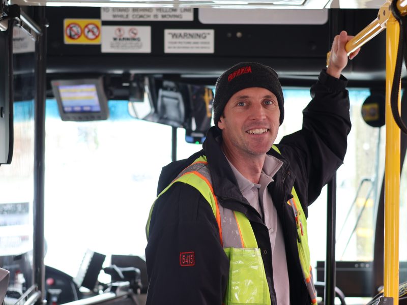 Man in high-visibility vest and beanie stands inside a bus, holding a handrail and smiling at the camera. Bus interior and dashboard are visible in the background.