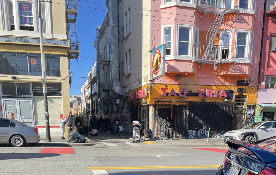 On a sunny day at the vibrant 16th St. Plaza, people gather near a colorful taqueria. Nearby buildings and parked cars add to the bustling street scene.