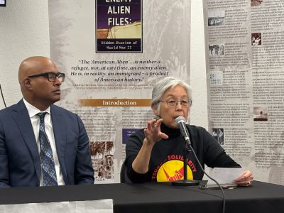 Two people seated at a table. The person on the right is speaking into a microphone, holding a piece of paper. Backdrop features text panels and a banner about "Enemy Alien Files.