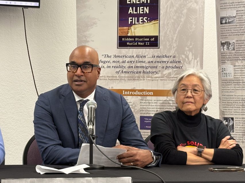 A man in a suit speaks into a microphone at a panel discussion. An older woman sits beside him. A poster about World War II is in the background.