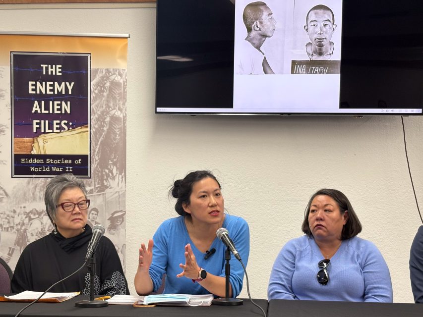 Three women seated at a table with microphones during a discussion. A poster titled "The Enemy Alien Files" and a black-and-white mugshot image are displayed in the background.