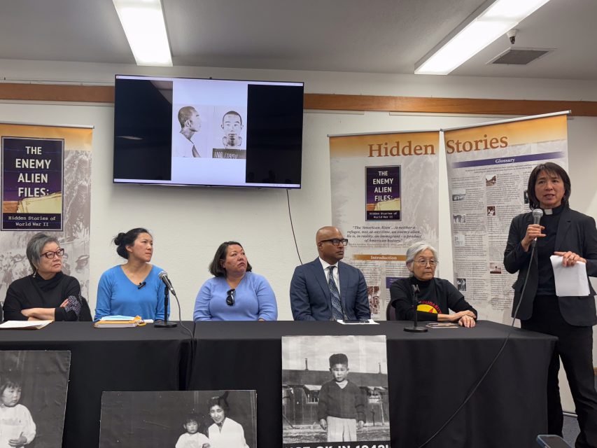 A group of six people sit or stand at a table with microphones in front of banners about historical topics. One person is speaking, and a black-and-white historical photo is projected on the wall.