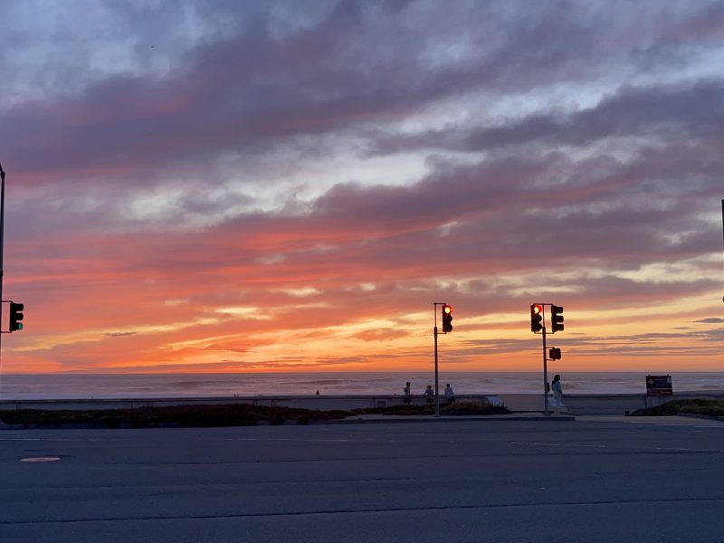 Traffic lights at a beachside intersection silhouetted against a vivid sunset sky with orange, pink, and purple clouds.