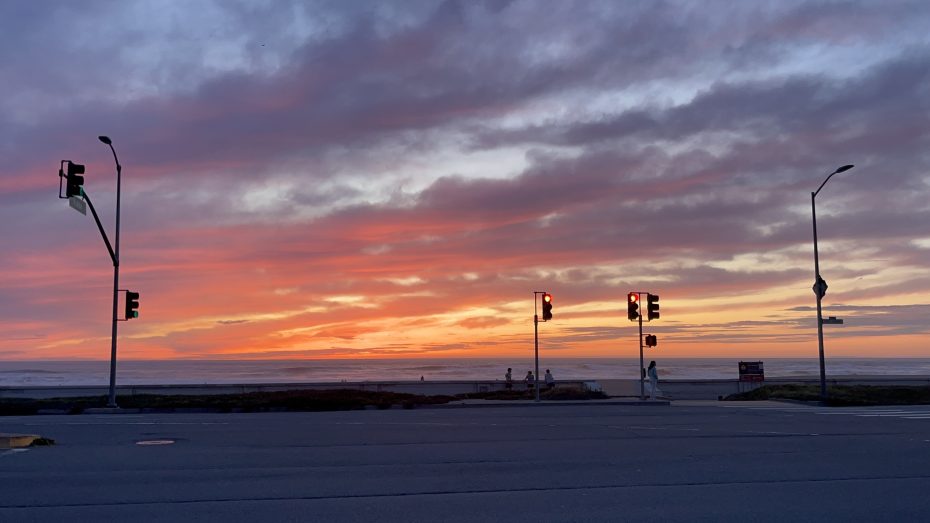 Traffic lights at a beachside intersection silhouetted against a vivid sunset sky with orange, pink, and purple clouds.