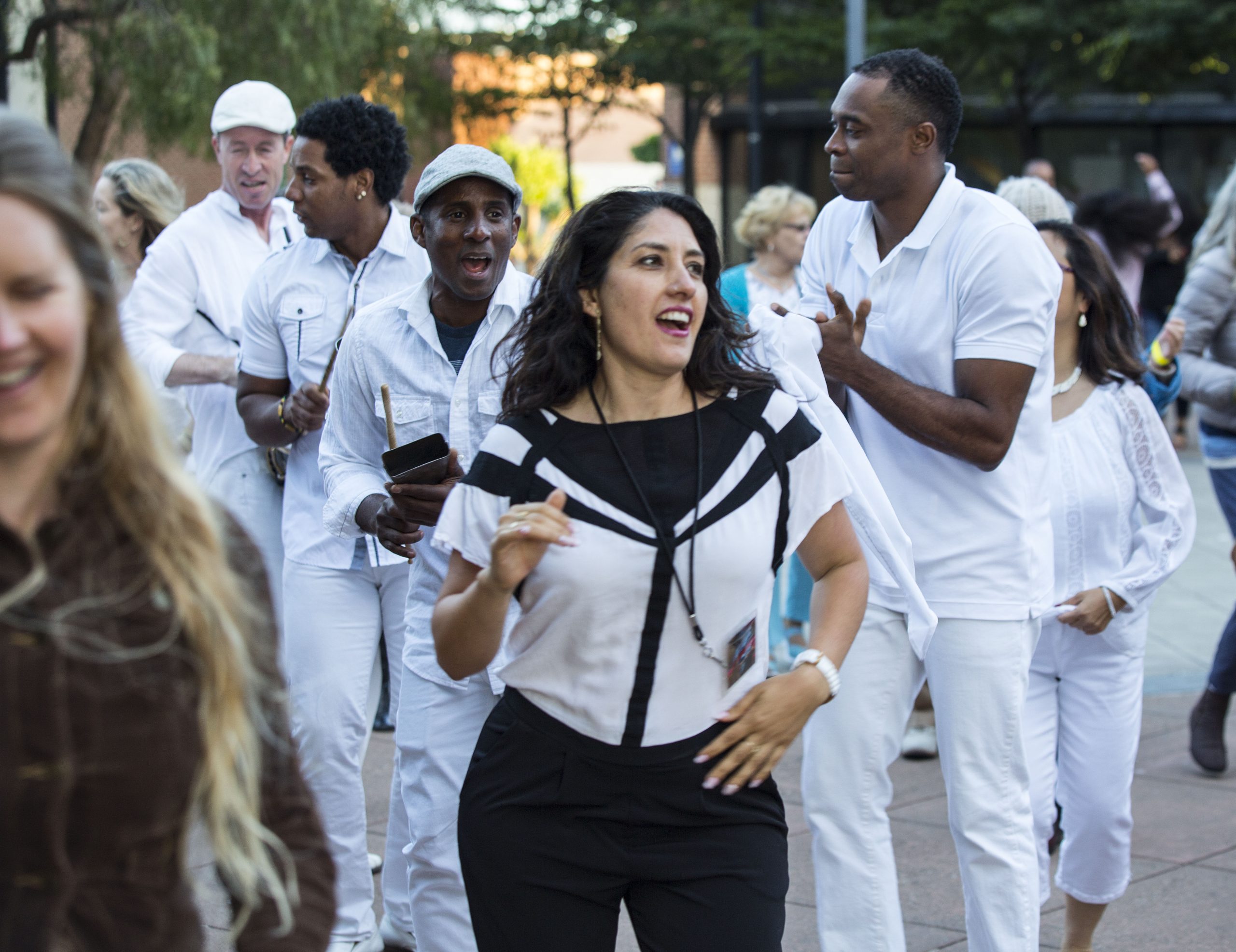A group of people dressed in white, outdoors, some clapping and smiling, as they walk together. Trees and buildings in the background.